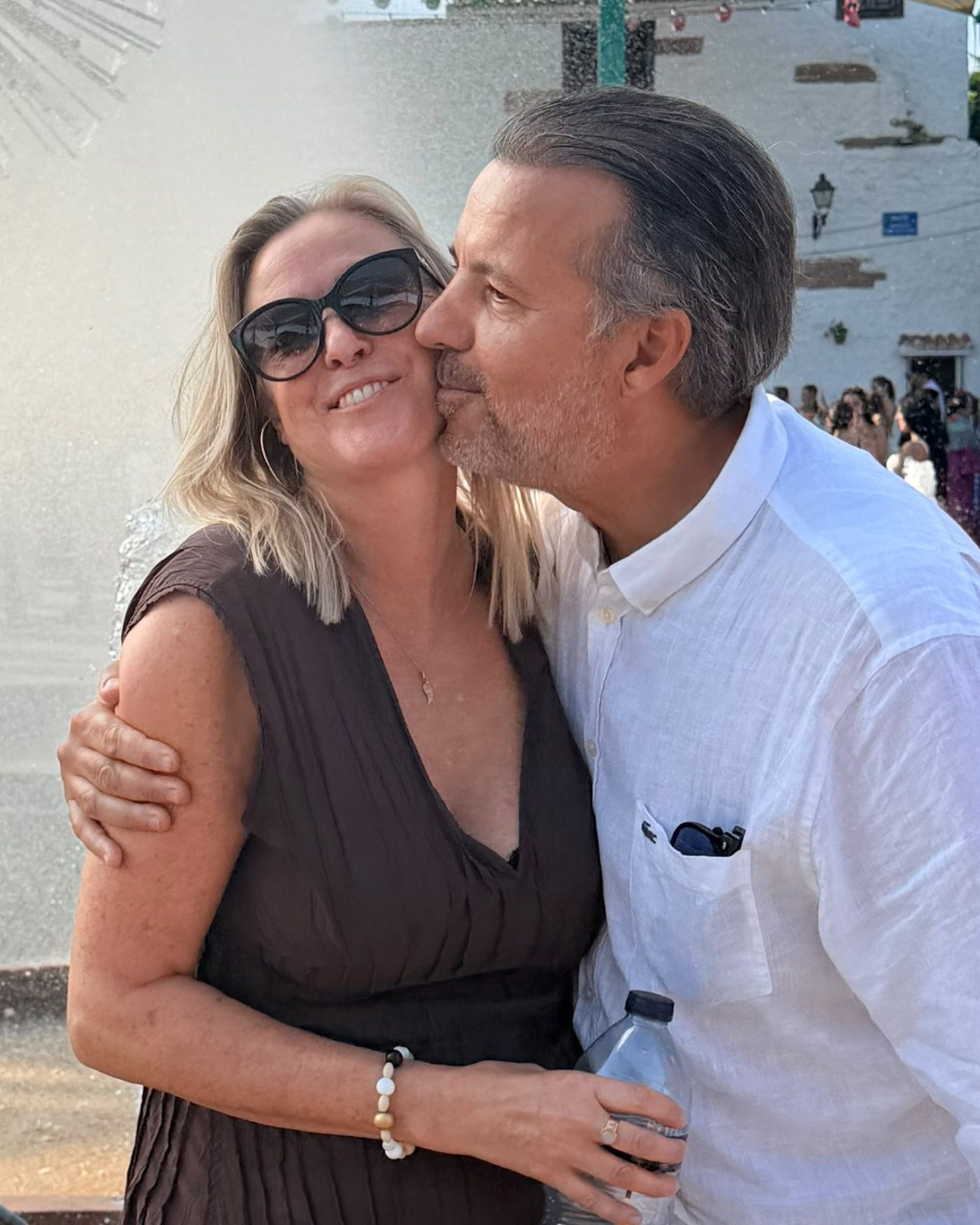 Sara Lee smiling as her partner kisses her cheek and holds her close, beside a fountain in a white Andalusian village.