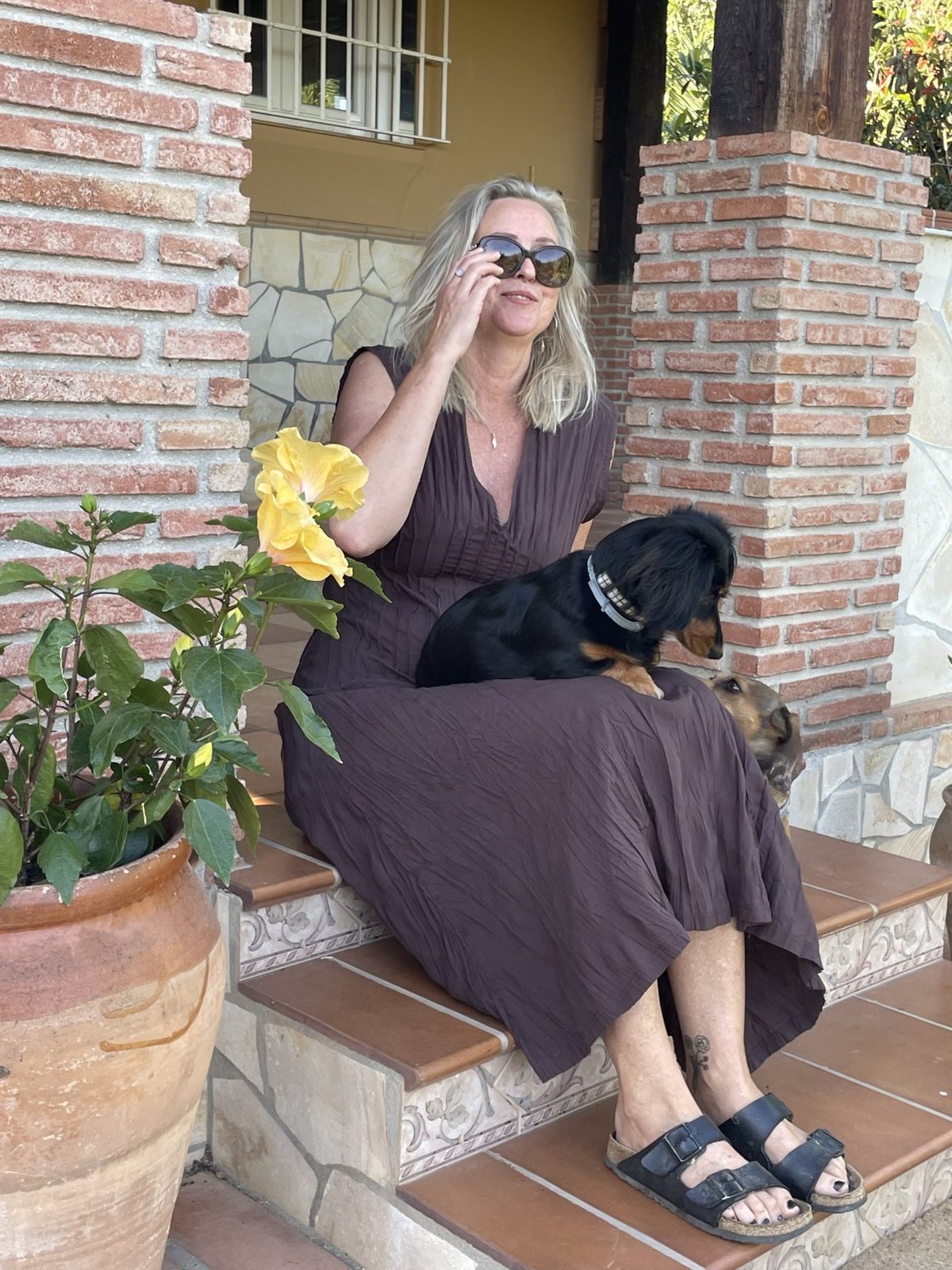 Sara Lee sitting on terracotta steps at Casa Eden in a long dress and sunglasses, a small dog on her lap and yellow hibiscus beside her.